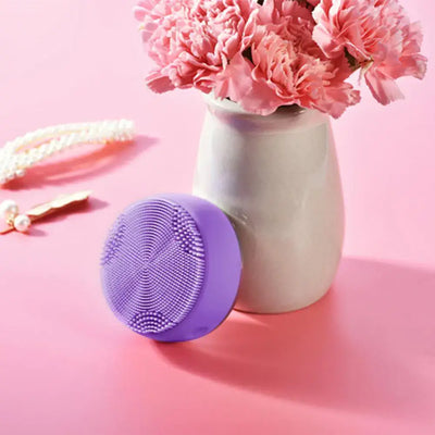 Electric Silicone Pore Cleaner and Heating Facial Wash next to a vase with pink flowers, on a pink surface.